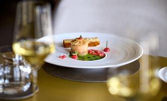 Ein kunstvoll angerichteter Teller mit feinem Gericht, umgeben von dekorativen Beeren und einem grünen Sauce. Im Hintergrund sind Weingläser zu sehen.