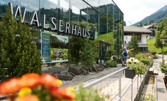 A modern building with a glass façade and the inscription "Walserhaus". In the foreground, colorful flowers are blooming, while in the background, the mountains and traditional buildings can be seen. | © Kleinwalsertal Tourismus | Oliver Farys