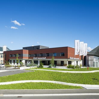 A modern building with a curved lawn area and a clear blue sky. In the background, trees and mountains are visible. | © Klinikverbund Allgäu