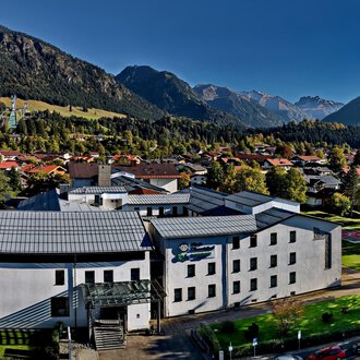 Ein modernes Gebäude umgeben von Bergen und Bäumen. Der Himmel ist klar und die Umgebung wirkt ruhig und einladend. | © Klinikverbund Allgäu