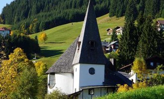 Ein charmantes, modernes Gebäude mit einem hohen spitzen Dach steht inmitten einer grünen Landschaft. Umgeben von Bäumen und sanften Hügeln strahlt die Szene Ruhe und Schönheit aus. | © Ev. Kirche Kleinwalsertal