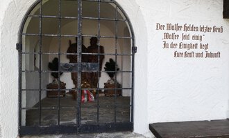 Ein historischer Eingang mit einem stabilen Gittertor. Im Inneren sind Statuen zu sehen, die mit Blumen geschmückt sind. | © Kleinwalsertal Tourismus | Nicole Lughammer