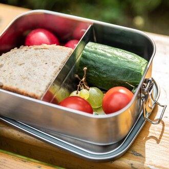 A lunchbox with fresh bread, cucumber, cherry tomatoes, grapes, and radishes. It is on a wooden surface in a natural setting. | © Kleinwalsertal Tourismus | Bastian Morell
