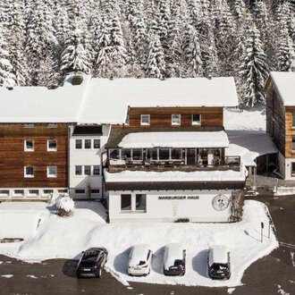 Eine schneebedeckte Landschaft mit mehreren Gebäuden im Chalet-Stil. Umgeben von hohen, schneebedeckten Tannenbäumen. | © Marburger Haus | Sweetchili
