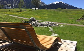 Eine Liege aus Holz steht auf einer Terrasse mit Blick auf grüne Wiesen und schneebedeckte Berge im Hintergrund. Der Himmel ist klar und die Landschaft wirkt idyllisch. | © Kleinwalsertal Tourismus | Sarina Berchtold