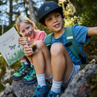 Two children are sitting on a stone in nature. One of the children is holding a book, while the other has a glass with something in their hand.