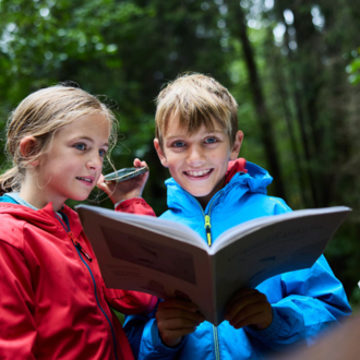 Zwei Kinder stehen im Wald und lesen ein Buch. Sie tragen bunte Regenjacken und lachen dabei.