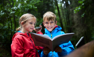 Zwei Kinder stehen im Wald und lesen ein Buch. Sie tragen bunte Regenjacken und lachen dabei.