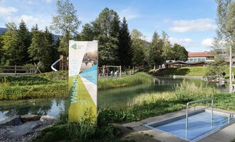 A small swimming pool in a green meadow, surrounded by trees. In the background, playground equipment and a building can be seen. | © Kleinwalsertal Tourismus | Veronika Senn