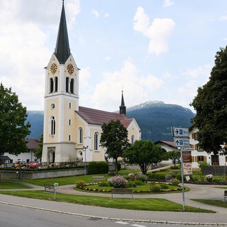 A quiet street in a charming village with a tall church in the background. Surrounded by trees and mountains, the scene radiates tranquility. | © Kleinwalsertal Tourismus | Oliver Farys