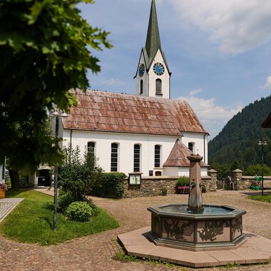 Ein malerischer Dorfplatz mit einem Brunnen im Vordergrund und traditionellen Häusern. Im Hintergrund ist eine Kirche mit einem hohen Turm und einem bewölkten Himmel zu sehen. | © Kleinwalsertal Tourismus | Oliver Farys