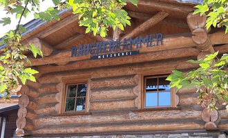 A cozy wooden house with windows and a decorative sign. Surrounded by green leaves and trees. | © Kleinwalsertal Tourismus | Veronika Senn