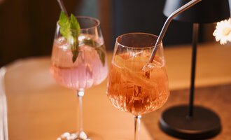 Zwei elegante Cocktails stehen auf einem Holztisch. Einer ist rosa mit Minze, der andere hat eine orange Farbe und einen Strohhalm.