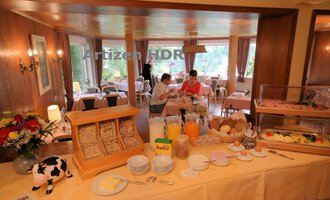 An inviting breakfast room with a buffet full of food and drinks. In the background, two people are sitting at a table and enjoying their breakfast. | © Hotel Bellevue | Werbewind GmbH