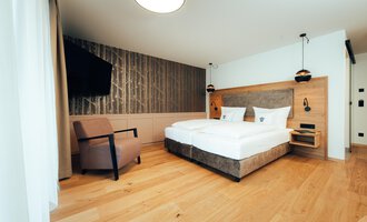 Ein modernes Hotelzimmer mit einem großen Doppelbett und einem gemütlichen Sessel. Die Wände sind in neutralen Tönen gehalten und sorgen für eine entspannte Atmosphäre.