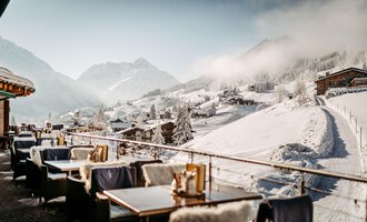 Ein schöner Blick auf eine verschneite Berglandschaft mit gemütlichen Tischen auf einer Terrasse. Die Atmosphäre ist ruhig und einladend, umgeben von schneebedeckten Hügeln und Bergen.