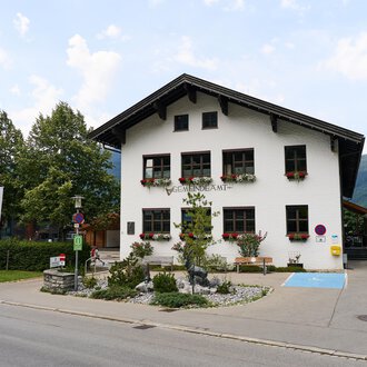 A charming, traditional building with a hipped roof. It is surrounded by trees and has blooming window boxes. | © Kleinwalsertal Tourismus | Oliver Farys