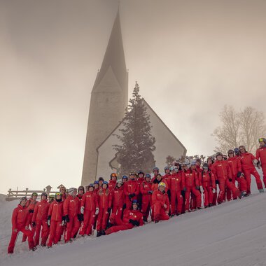 A group of people in red ski suits is standing in the snow in front of a church. The landscape is wintry and surrounded by fog. | © Skischule Mittelberg | Karl Schuster