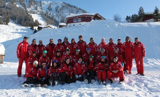 A group of skiers in red suits is posing in the snow. In the background, snow-covered mountains and cabins can be seen. | © Skischule Riezlern GmbH | Frank Felder