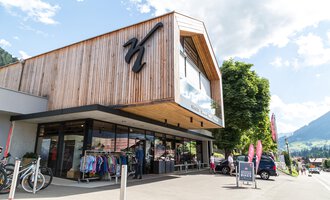 A modern shop with a wooden facade and large windows. In the foreground, clothing and bicycles are displayed, surrounded by a beautiful mountain landscape. | © Sport Kessler GmbH