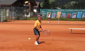 Ein Mann spielt Tennis auf einem roten Platz mit Blick auf die Berge. Im Hintergrund sind weitere Spieler und eine Tenniswand zu sehen. | © Sportverein Kleinwalsertal