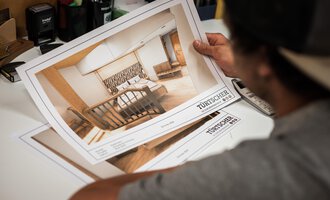 A person is looking at plans for interior design. The plans show a modern, inviting room with a bed and a comfortable seating area. | © Türtscher Die Schreinermanufaktur