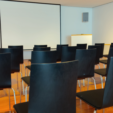 A room with black chairs and a projection screen. The floor is made of wood and the walls are bright.