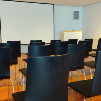 A room with black chairs and a projection screen. The floor is made of wood and the walls are bright.