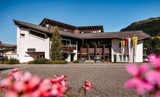 Ein modernes Gebäude mit einer eleganten Fassade und umgeben von schönen Blumen. Im Hintergrund sind Berge und ein klarer Himmel sichtbar. | © Walser Raiffeisen Bank AG