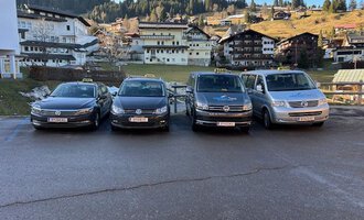 Vier Taxis stehen in einer Reihe vor einer malerischen Berglandschaft. Im Hintergrund sind traditionelle alpine Gebäude und bewaldete Hügel zu sehen. | © Walser Taxi | Josef Düringer