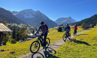 Drei Radfahrer fahren auf einem Weg durch eine grüne Landschaft mit Bergen im Hintergrund. Der Himmel ist klar und blau. | © WalserBike Tours | Christian Gutermuth