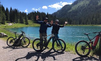 Two cyclists stand by the shore of a clear lake, smiling happily at the camera. In the background, there are green mountains and a clear blue sky. | © WalserBike Tours | Christian Gutermuth
