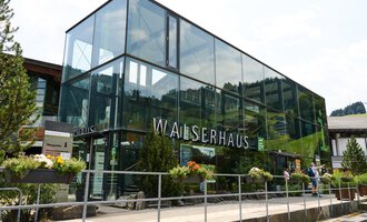 Ein modernes Gebäude mit großen Glasfenstern, umgeben von grünen Pflanzen. Es trägt die Beschriftung "WALSERHAUS". | © Kleinwalsertal Tourismus | Oliver Farys