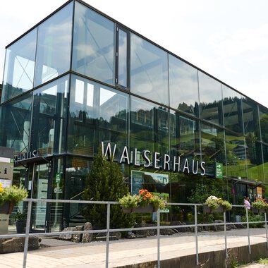 A modern building with large glass windows, surrounded by green plants. It bears the inscription "WALSERHAUS". | © Kleinwalsertal Tourismus | Oliver Farys