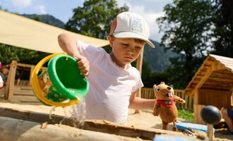 A boy in the sandbox is pouring sand from a bucket. Next to him, he is holding a stuffed toy. | © Kleinwalsertal Tourismus | Oliver Farys