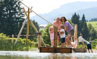 A group of children and adults is standing on a wooden bridge over a calm pond. In the background, trees and mountains can be seen. | © Kleinwalsertal Tourismus | Oliver Farys