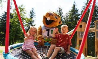 A cheerful playground with two children and a mascot. They are sitting in a large scoop seesaw and enjoying the sun. | © Kleinwalsertal Tourismus | Oliver Farys