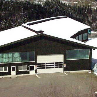 A large, modern building with a black facade and a snow-covered roof. Surrounded by snowy trees and a peaceful landscape. | © Gemeinde Mittelberg