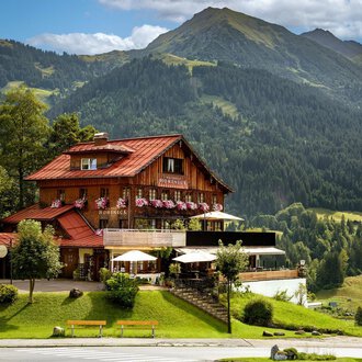 Ein rustikales Chalet in den Bergen mit einem roten Dach und vielen Blumen. Die Umgebung ist grün und hügelig, umgeben von majestätischen Bergen.