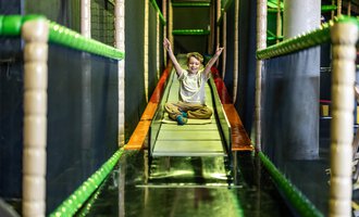 Ein fröhliches Kind sitzt auf einer Rutsche in einem Spielplatz. Die Umgebung ist bunt und einladend für Kinder. | © Wonniland Sonthofen | interSPA Gruppe