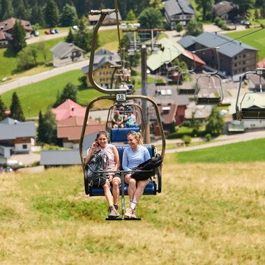 Two women are sitting in a chair lift and riding up a mountain meadow. In the background, green hills and a small village with houses can be seen. | © Kleinwalsertal Tourismus | Oliver Farys