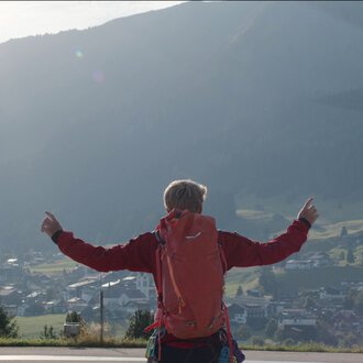 Eine Person mit rotem Rucksack steht auf einer Wiese und zeigt nach oben, während ein Hubschrauber im Flug über der Landschaft fliegt. | © Kleinwalsertal Tourismus eGen