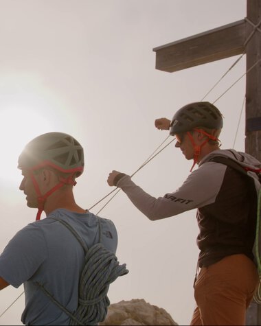 Zwei Kletterer befestigen sich in den Bergen an einem Seil, mit einem Holzmast im Hintergrund und der Sonne im Himmel. | © Kleinwalsertal Tourismus eGen