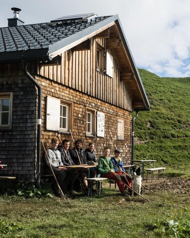 A group of people sitting outside an old wooden house in a lush, mountainous landscape. | © Kleinwalsertal Tourismus eGen • Photographer: Andre Tappe