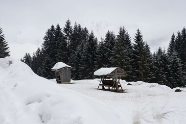 Snowy landscape with wooden huts and a shelter in a forested winter scene. | © Kleinwalsertal Tourismus eGen • Photographer: Andre Tappe
