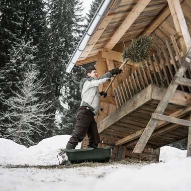 Junge Person wirft Heu oder Zweige von einem Balkon eines Holzhauses in einer verschneiten Waldumgebung. | © Kleinwalsertal Tourismus eGen • Fotograf: Andre Tappe