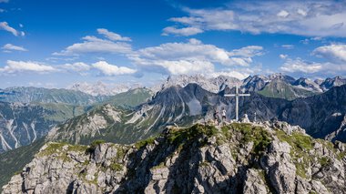 Berggipfel mit Gipfelkreuz und Wanderern, oberhalb der Alpen, bei schönem Wetter, mit blauer Himmel und Wolken. | © Kleinwalsertal Tourismus eGen • Fotograf: Bastian Morell