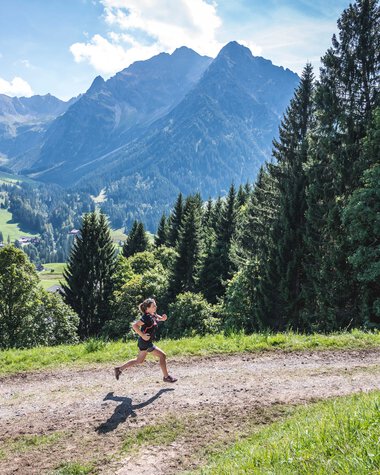 Jemand läuft auf einem Wanderweg in einer bergigen, grünen Landschaft mit hohen Bergen im Hintergrund. | © Kleinwalsertal Tourismus eGen • Fotograf: Dominik Berchtold