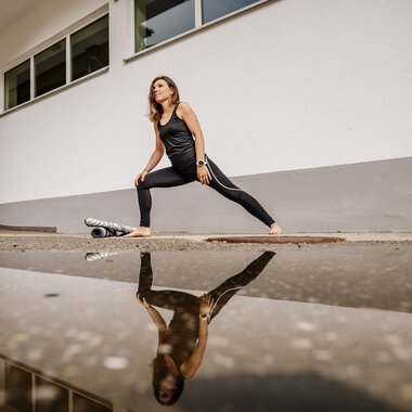 Frau macht Yoga-Übung im Outdoor-Bereich, mit Reflexion im Wasser, vor weißem Gebäude. | © Kleinwalsertal Tourismus eGen • Fotograf: Dominik Berchtold