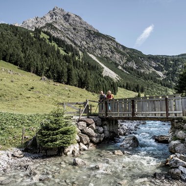 Zwei Personen auf einer Holzbrücke über einen Bach in einer alpinen Landschaft mit Bergen im Hintergrund. | © Kleinwalsertal Tourismus eGen • Fotograf: Dominik Berchtold